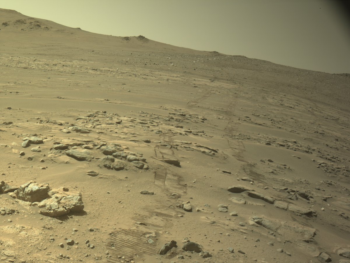 Rover tracks crossing a pale, rocky Martian slope toward a distant ridge beneath a hazy sky.