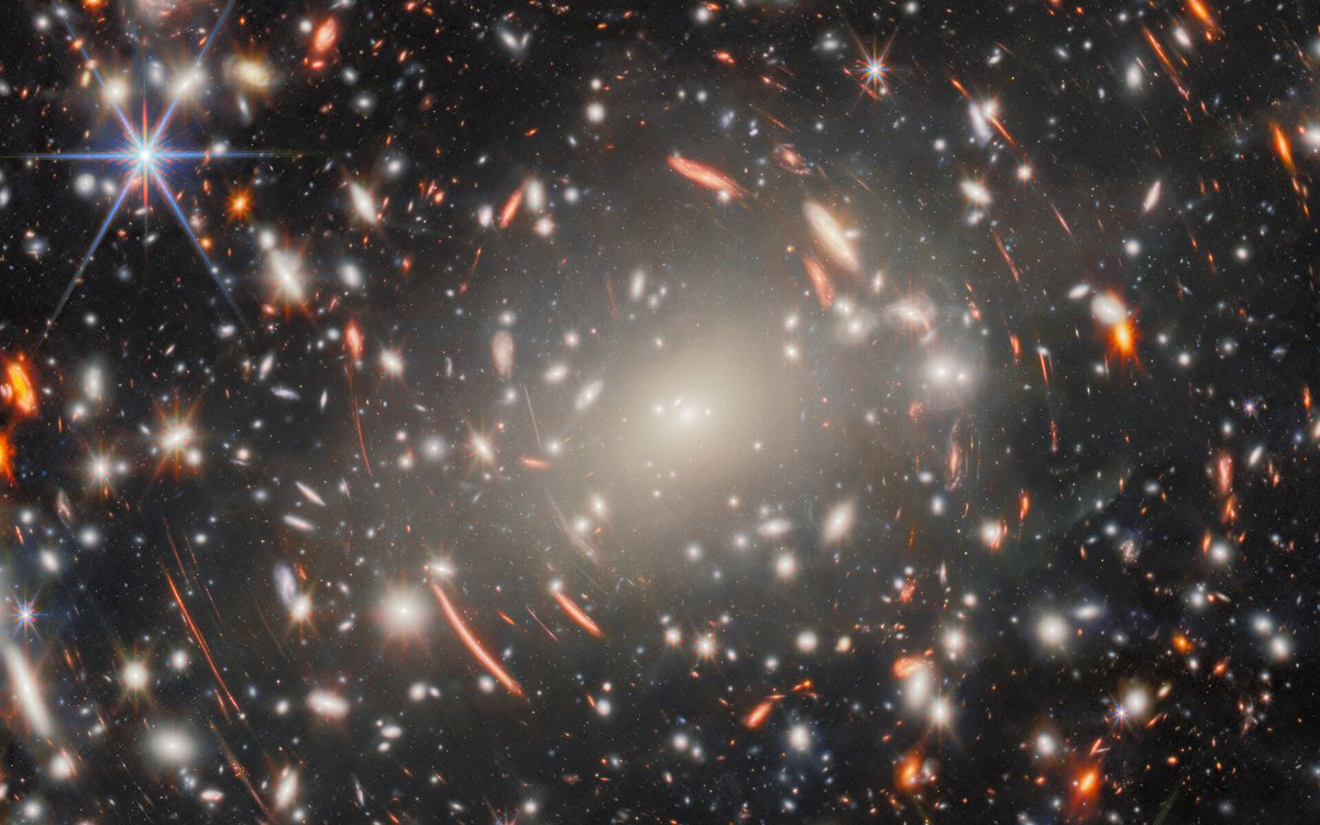 Hundreds of galaxies and stars surrounding a bright central galaxy cluster, with elongated red arcs scattered across the scene.