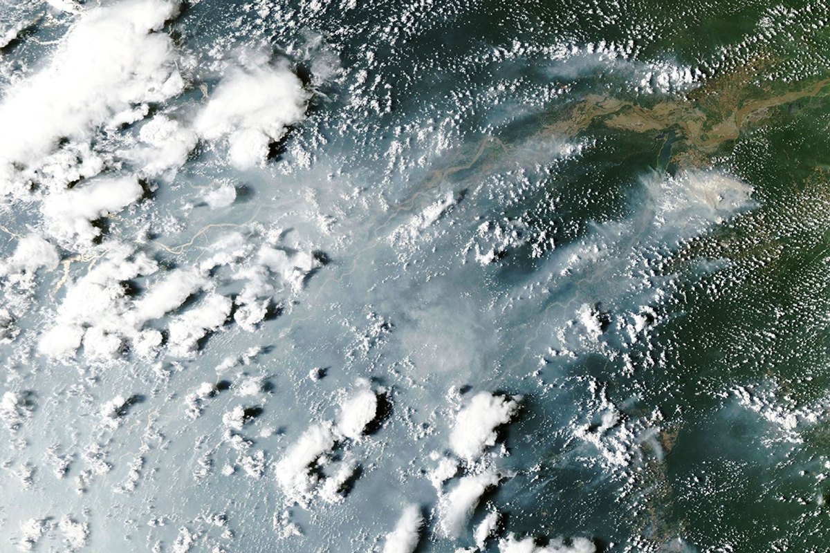 Satellite photograph of gray smoke and white clouds spreading across the Amazon River and surrounding green rainforest.