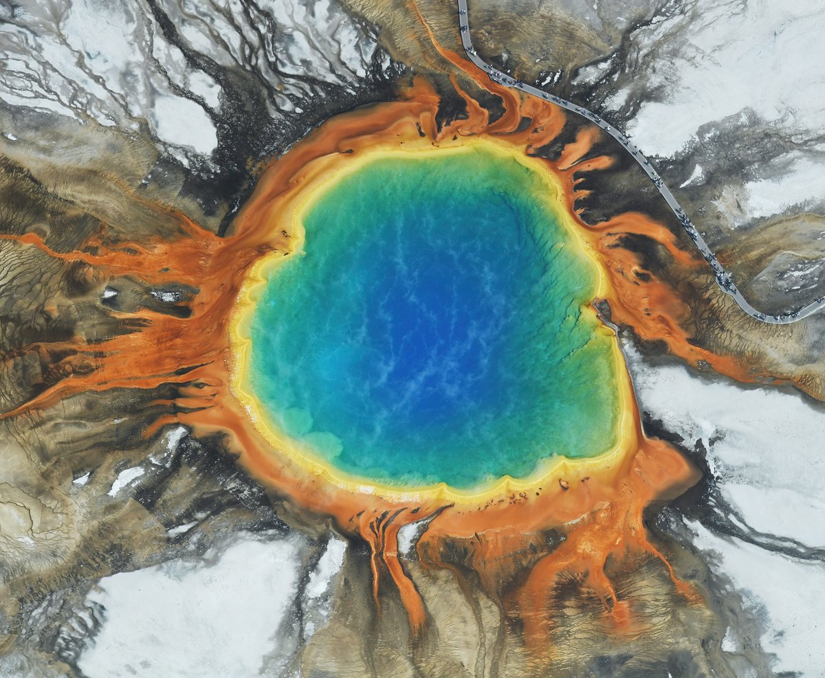 Aerial view of a steaming circular blue hot spring surrounded by yellow, orange and green bands.