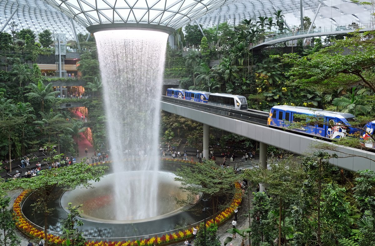A towering indoor waterfall plunging from a glass roof into a circular basin surrounded by tropical greenery.