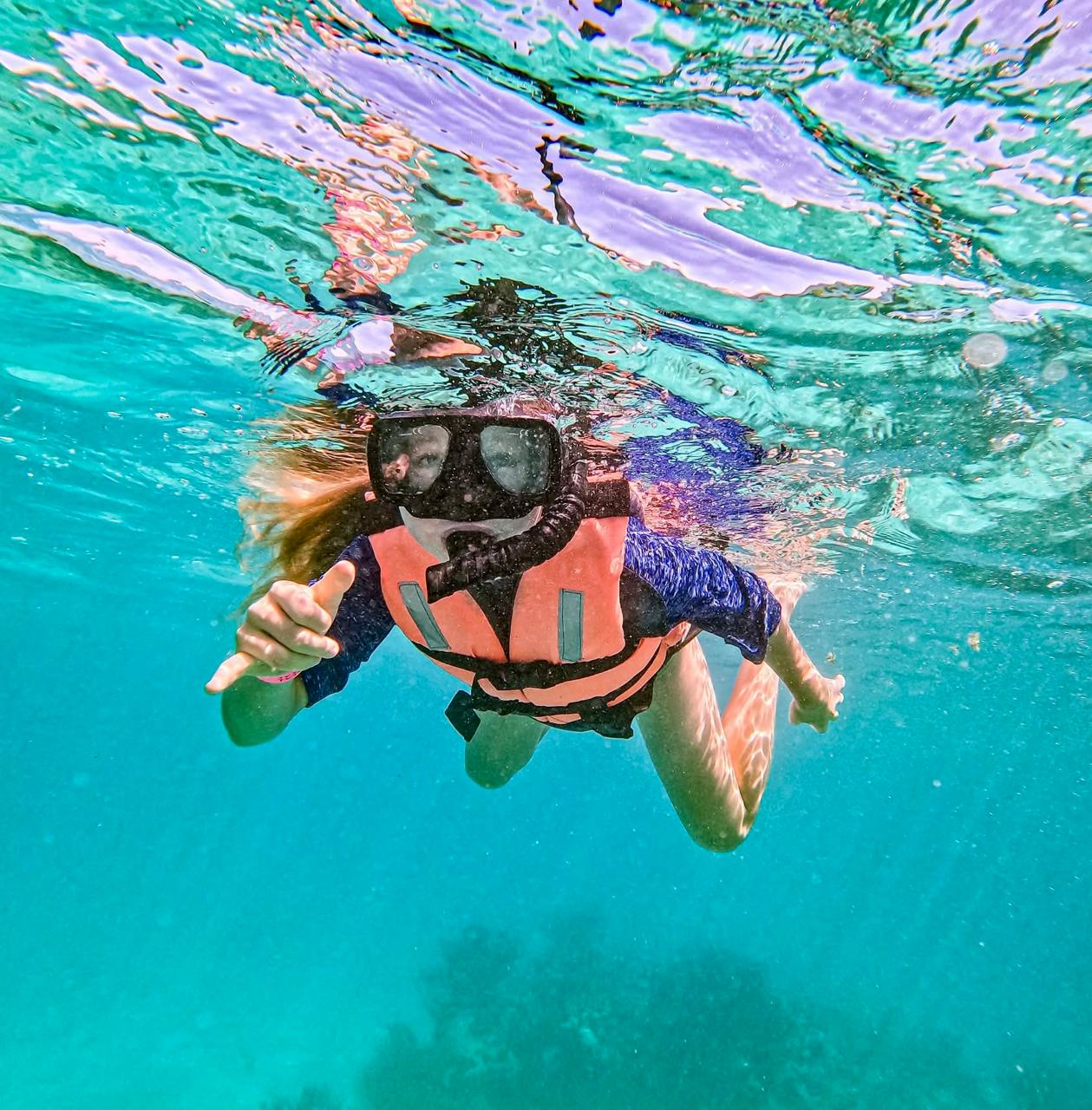 Aimee snorkelling underwater in a life vest, making a shaka sign