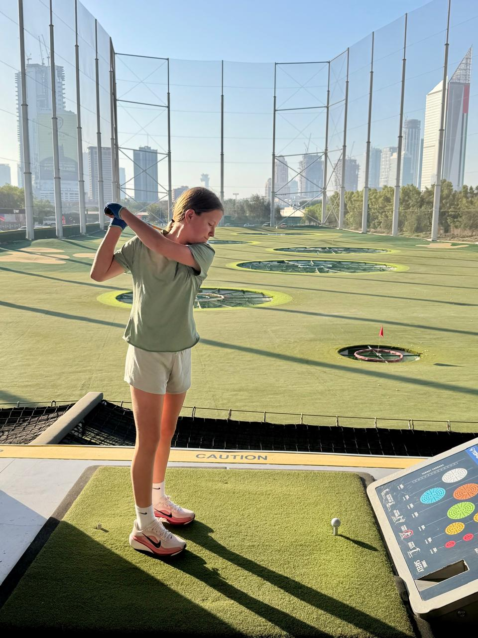 Aimee mid-swing at a driving range with the Dubai skyline behind