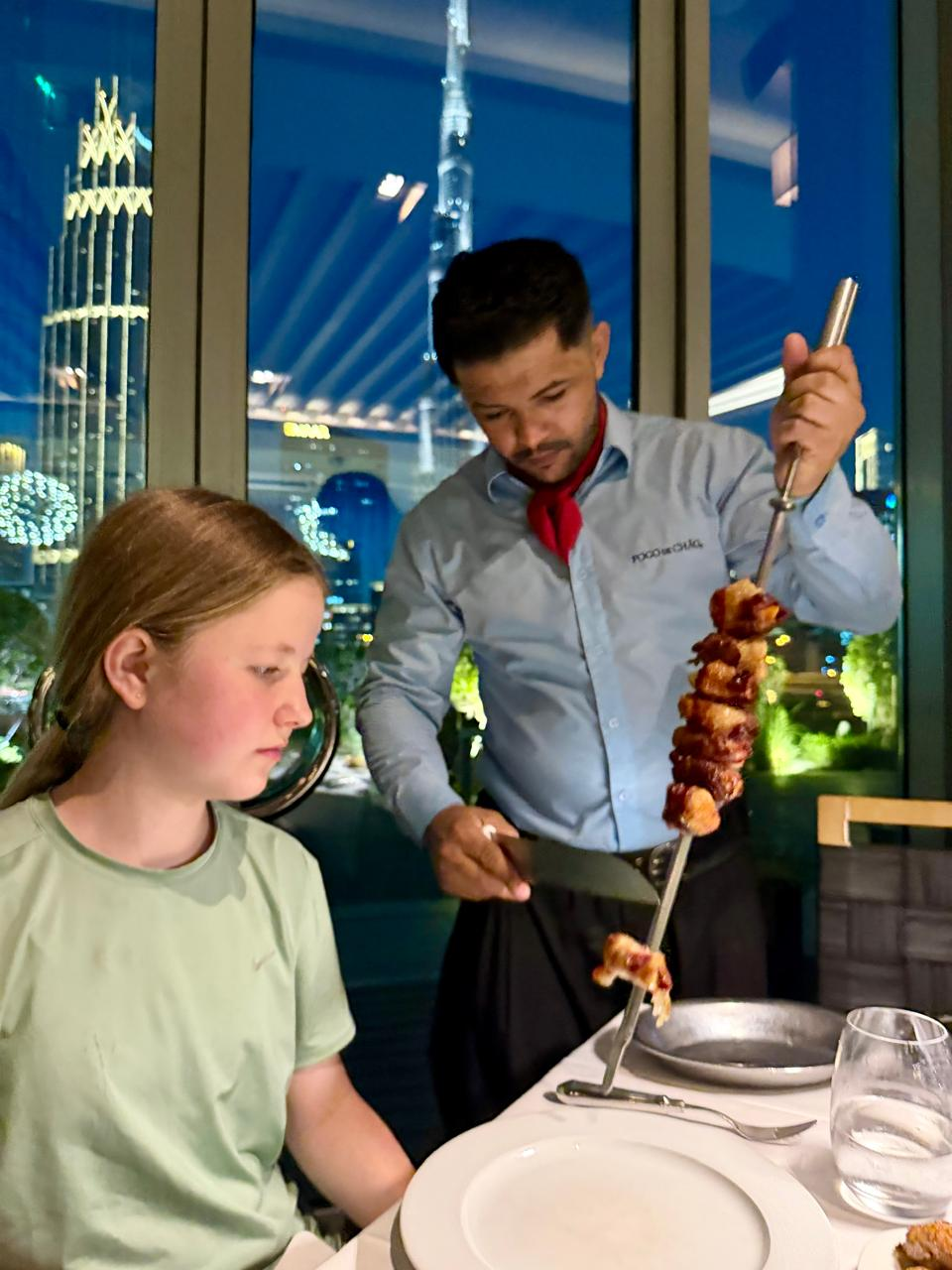 A waiter carves meat from a skewer beside Aimee, with the Burj Khalifa lit up behind them