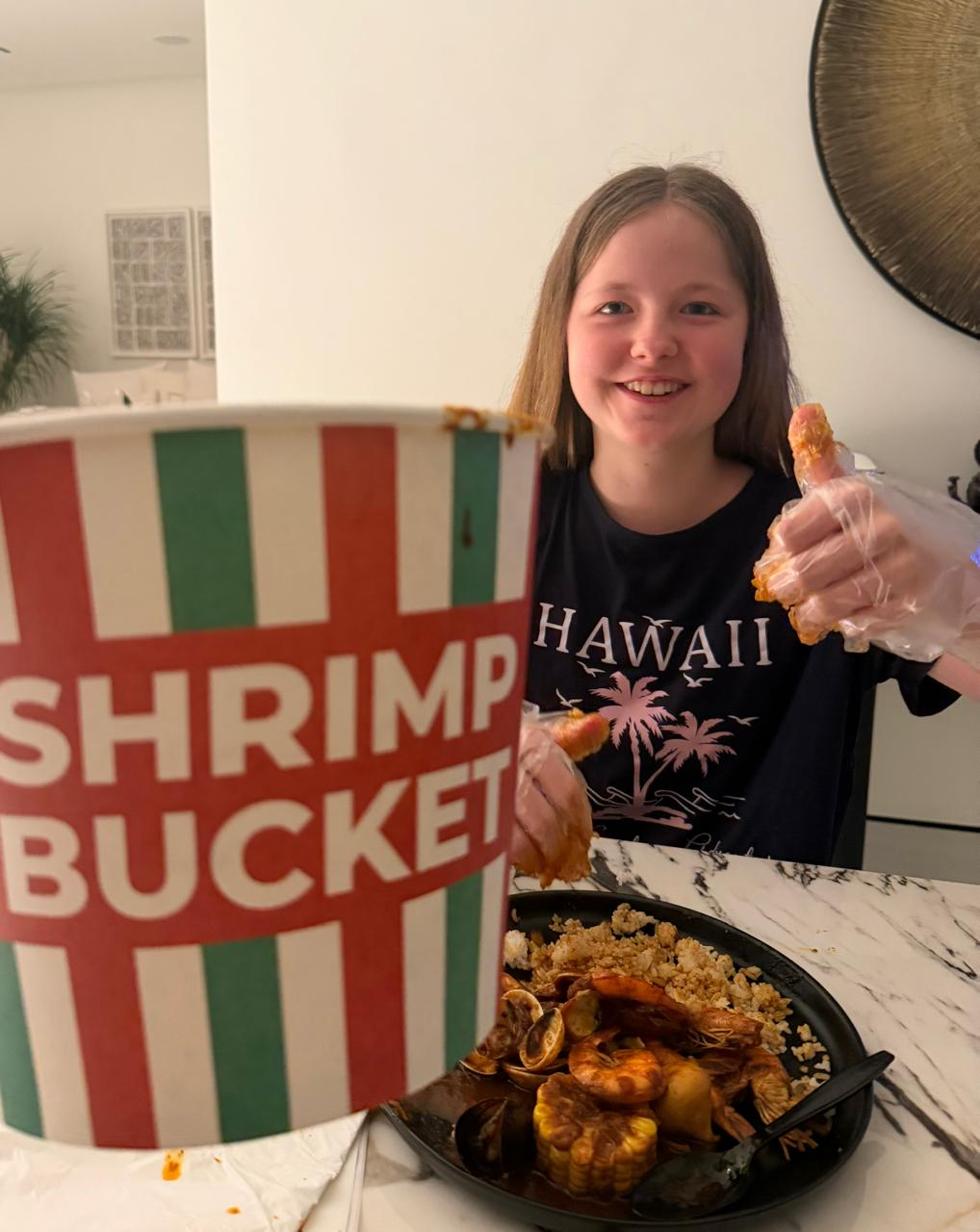 Aimee at home giving a thumbs up with a shrimp, behind a Shrimp Bucket delivery tub