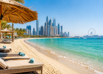 Turquoise water, soft sand and beachfront loungers with the Dubai Marina skyline and Ain Dubai in the distance.