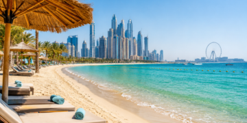 Turquoise water, soft sand and beachfront loungers with the Dubai Marina skyline and Ain Dubai in the distance.
