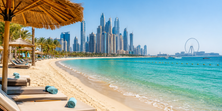 Turquoise water, soft sand and beachfront loungers with the Dubai Marina skyline and Ain Dubai in the distance.